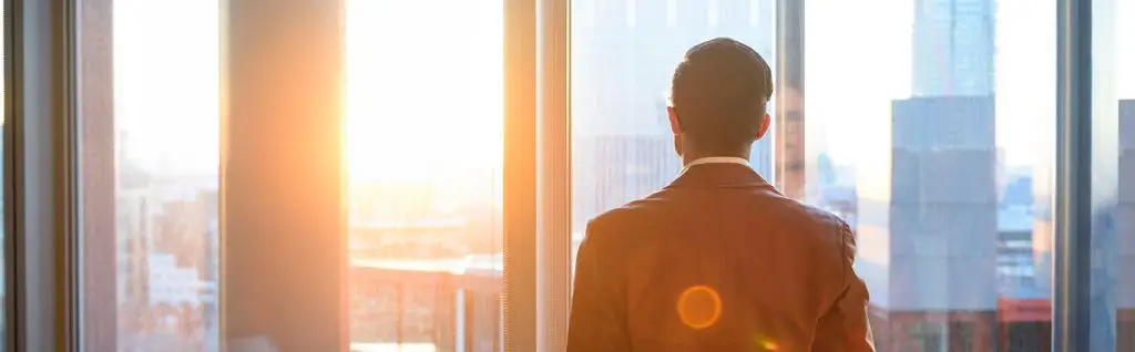 a man in a brown blazer looking out the office window during sunset
